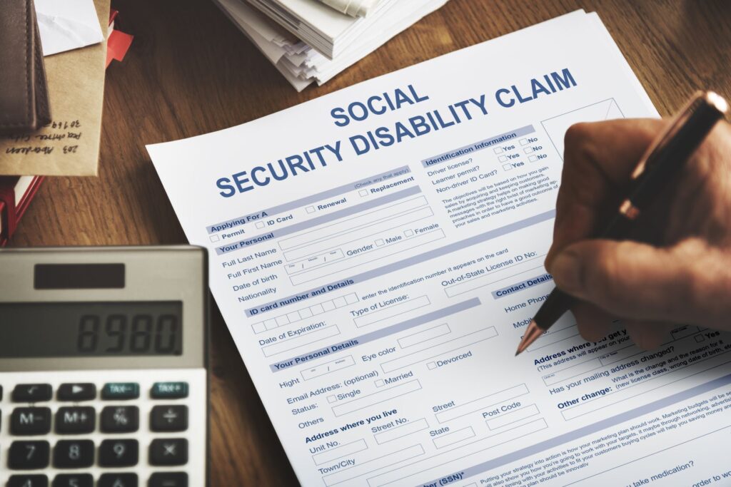 A person filling out a Social Security Disability Claim form on a wooden table, with a calculator and papers nearby.
